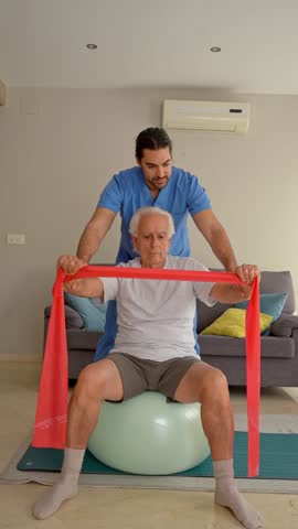 Vertical video of Physiotherapist assisting senior man with resistance band exercises on fitness ball during home visit