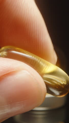 Vertical video. Fingers Squeezing an Omega 3 Capsule to Show Its Softness and Elasticity in a Macro Close-Up with a Gel Capsule Bottle in the Background Under Warm Lighting and a Dark Background