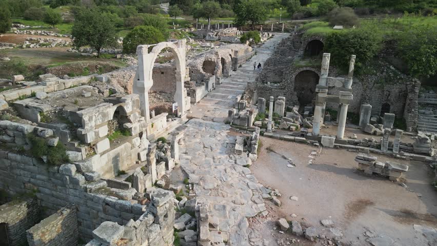 View from above of stone roads and arches in Ephesus, Turkey. Ancient Roman masonry preserved. Bright spring greenery surrounds ruins. Tourists walk the historic marble streets