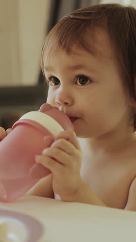 Vertical screen: Happy toddler drinking water from a pink cup or bottle with a straw surrounded by toys, sitting at a table in a bright, warm, light room on a sunny day