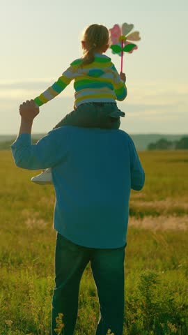 Child has pinwheel toy in his hand, daughter sitting on dad's shoulders. Happy family walk, kid girl dad outdoors. Dad kid girl are walking playing together, holding hands in park. People sunny park