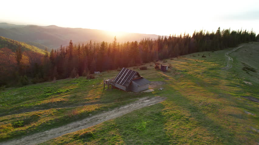 Aerial shot of serene sunset over hilly landscape in Ukraine. Rustic wooden cabin and dirt path nestled among rolling green hills, with forest and mountains bathed in warm, golden light.