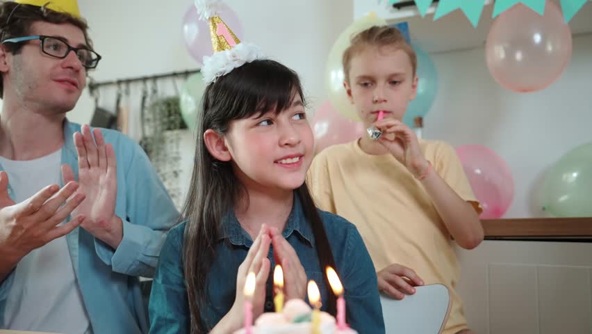 Happy cute child making a wish and blowing birthday cake candle surrounded by family wearing party hat celebrate in special occasion. Closeup of girl blow candle while family clapping hands. Pedagogy.