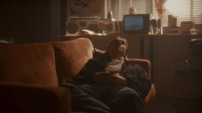 Young pensive man resting on sofa in cozy living room filled with soft golden sunlight, with books, vintage analog TV and boombox in background. Zoom shot - Powered by Shutterstock - Get 15% off with code: PIKWIZARD15
