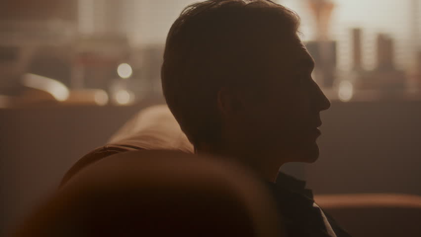 Young pensive man leaning back on a sofa in warmly lit living room, looking up deep in thought. Close-up view