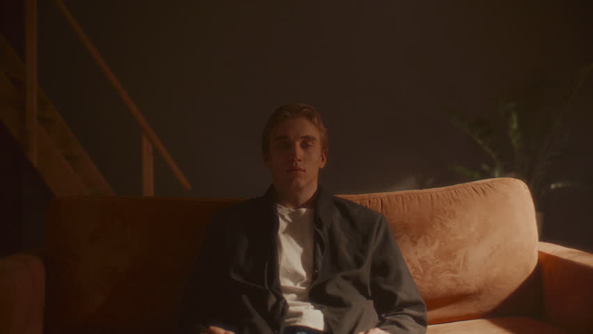 Young man sitting still on comfortable sofa, looking directly into camera in warmly lit minimalist room. Zoom shot, video portrait