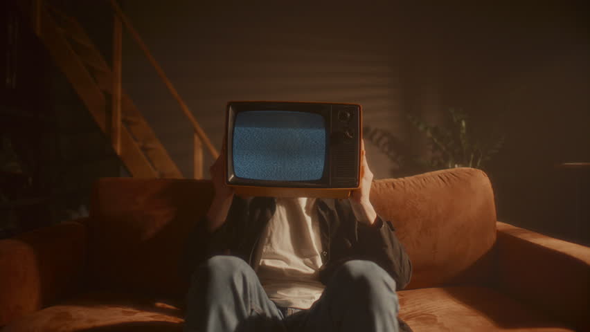 Young man sitting on sofa in warmly lit living room, holding vintage analog TV with static screen in front of his face. Zoom shot
