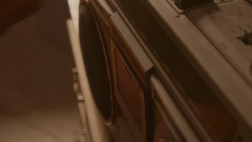 Close-up view of hand inserting audio cassette, then pressing play button on control panel of vintage boombox in warmly lit room