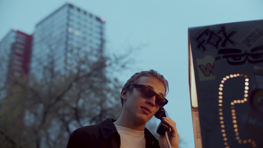 Young stylish man in sunglasses standing at decorated phone booth, having conversation during early evening in city with urban buildings and trees in background. Low angle view