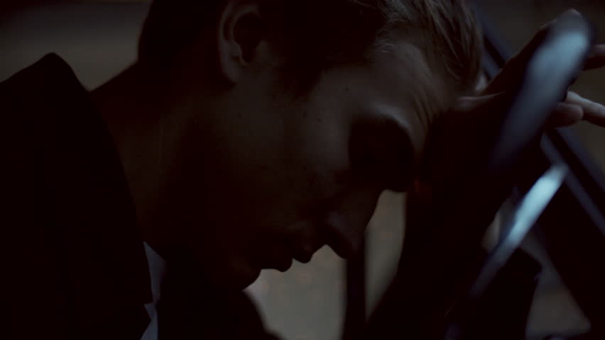 Young pensive man resting his head on steering wheel, then tilting head up and looking through windshield while sitting in car at night illuminated by multicolored lights of police car outside