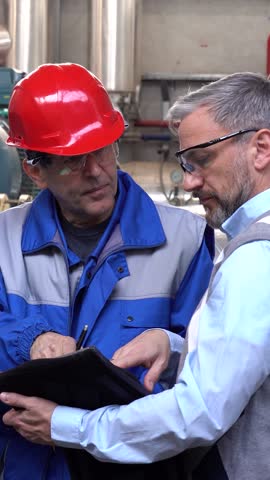Maintenance Engineer Uses Digital Tablet and Talking to Worker in Protective Workwear. Supervisor with gray hair in communication with maintenance technician discuss production process in a factory.