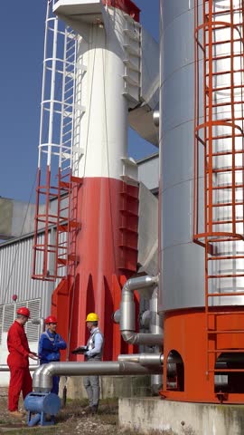 Two Workers and Production Manager on Meeting in front of Industrial Chimney and Storage Tanks. Middle-aged Supervisor using digital tablet in discussion with two workers in protective workwear.