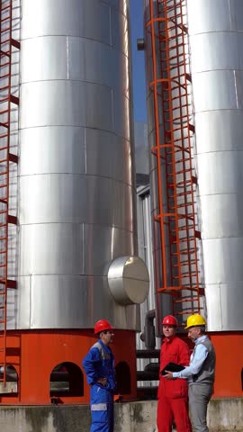 Oil Refinery Workers and Production Manager on Meeting in front of Industrial Storage Tanks. Middle-aged Supervisor using tablet in discussion with two workers in protective workwear. Energy Industry.