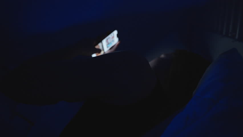 Woman on Bed at Night Using Smartphone with Face Lit by Screen Light in Low Angle Shot, Digital Addiction Concept