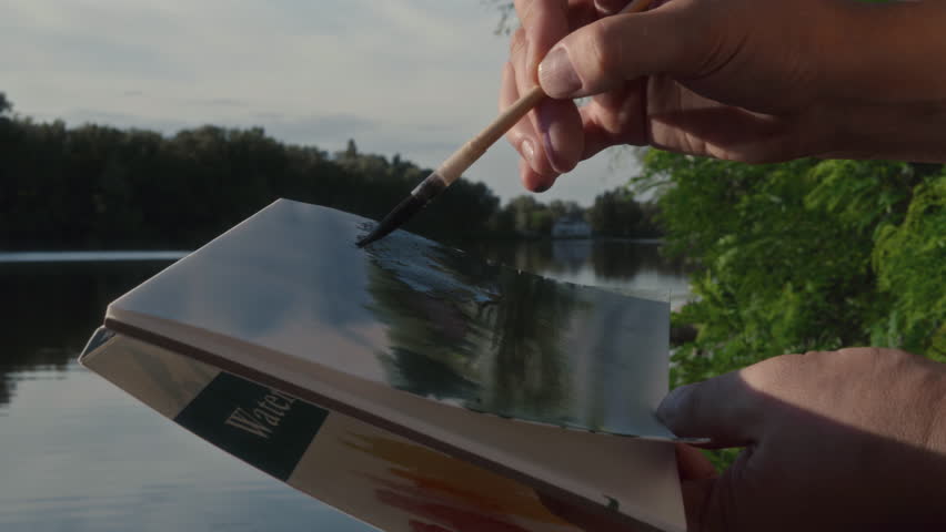 Close-up, female artist painting landscape with watercolor in album on river bank