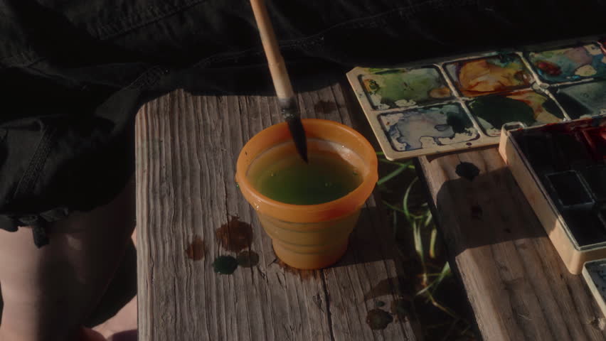 Close-up, the hand of a female artist washing a brush in a cup of water on a wooden pier