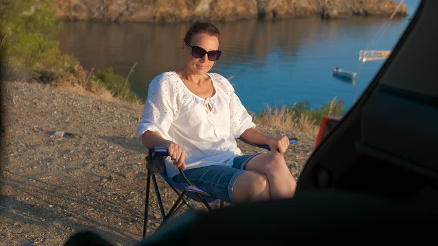 Woman unfolding camping chair by the lake. Experienced traveler unpacking lightweight folding chair from vehicle trunk, preparing for peaceful lakeside relaxation during scenic summer getaway