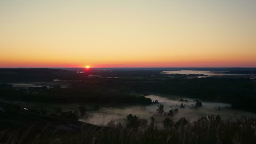 Foggy morning over plain, river floodplain meadow near rural village, landscape from birds eye view. Sunrise on summer morning over river, beautiful natural landscape. Sun is seen above horizon, sky