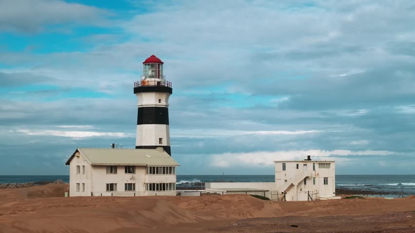Black and white striped lighthouse overlooks ocean from sandy beach, with small building nearby. Sky is filled with soft clouds. Cape Recife lighthouse in South Africa at dawn