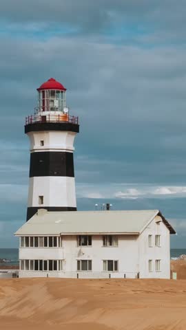 Black and white striped lighthouse overlooks ocean from sandy beach, with small building nearby. Sky is filled with soft clouds. Timelapse of Cape Recife lighthouse in South Africa at dawn