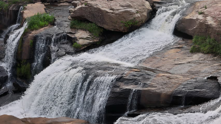 Small waterfall flows over rocks, creating ripples in clear water under bright sunlight at midday.