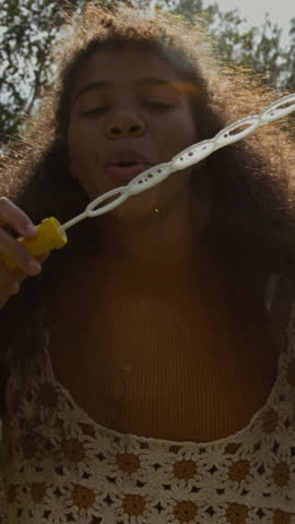 Vertical slowmo close-up with lens flare of happy teenage African American girl in stylish crochet top making soap bubbles with long wand, while relaxing outdoors in hot summer sun