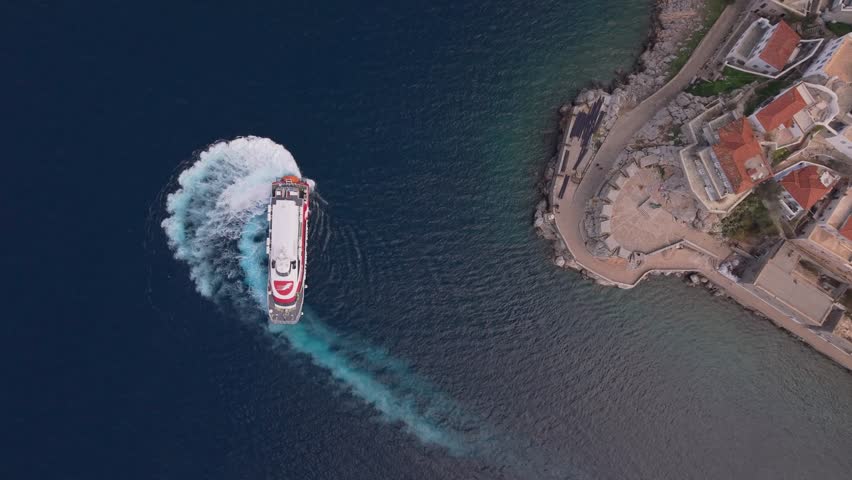 A top-down drone view of a ferry turning and leaving the port of Hydra, leaving behind a white wake in the blue Aegean Sea. A dynamic and cinematic maritime scene