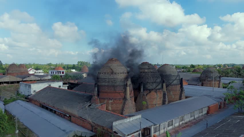 Dark fumes exhausting to blue sky with fluffy clouds from potteries kiln chimney, Mang Thít, Mekong delta, south Vietnam. Drone orbit factory view.