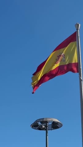 Vertical Slow motion of weathered Spanish flag fluttering from a pole, its edges frayed, threads loose, and fabric slightly torn. The red and yellow hues and national coat of arms against a wall