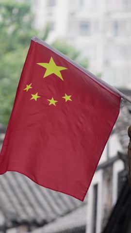 Vertical Slow motion of a Chinese flag with bright red and yellow colors is mounted on a tree, waving gently. Traditional architecture and greenery form background, scene rich in cultural significance