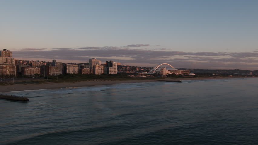 Dawn aerial over the sea from Moses Mabhida Stadium along the beachfront, capturing waves rolling onto the shore, the city skyline and the beachfront glowing in the sunlight.