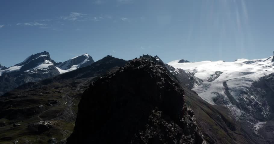 Drone orbiting tall mountain near Zermatt with hikers and summit cross, framed by snowy Alpine peaks, glaciers, and clear blue sky. A cinematic view of Swiss adventure and nature.