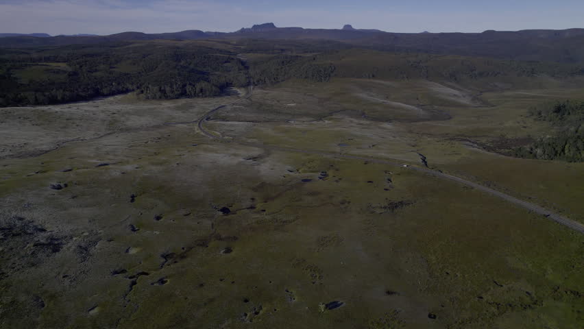 Drone captures a van driving through Tasmania’s frosty wilderness at dawn. Grassy plains, a winding river, and Cradle Mountain NP rise in the distance as the road leads into a forested horizon.