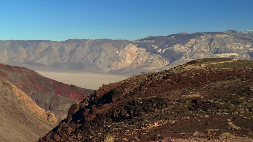 Static view of rugged rocky ridge in the foreground, transitioning into multicolored hills and a vast pale desert basin, framed by steep mountain slopes in Death Valley National Park, USA