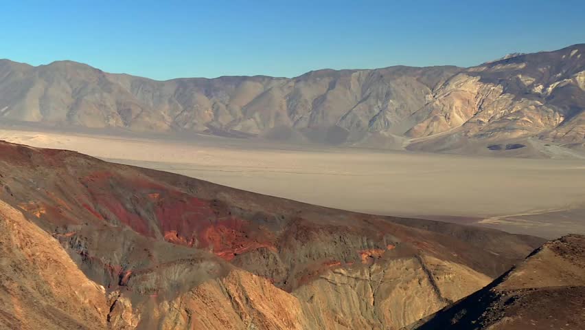 Vibrant reddish and gray ridges transitioning into the vast, flat valley floor, with rugged mountains rising in the background in Death Valley National Park, USA.