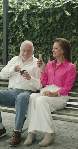 A sweet scene of a couple of grandparents enjoying ice cream together while sitting on a park bench on a sunny day.