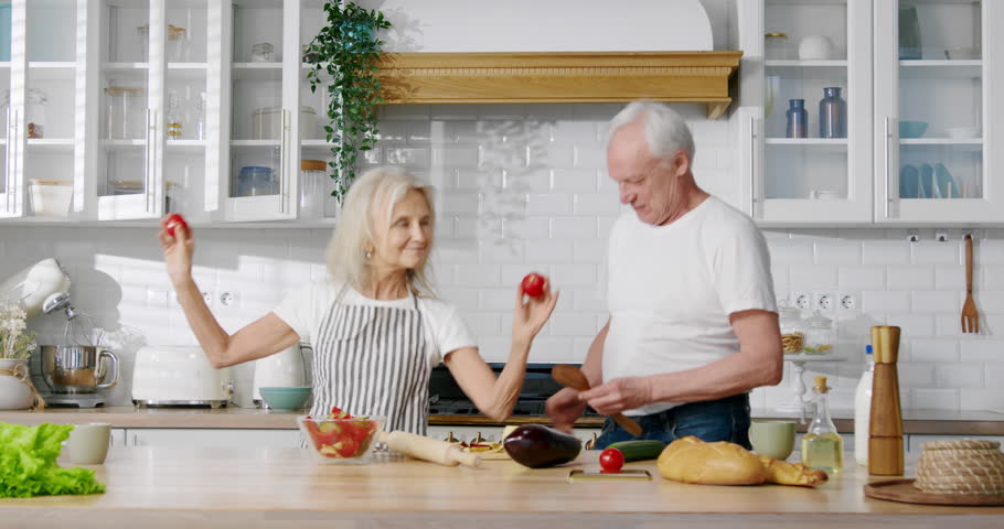 Aged playful married couple dancing in kitchen, portrait of funny grandparents. Elderly spouses listening to favorite song and moving by rhythm, joyful grandma and grandpa cooking dinner at home