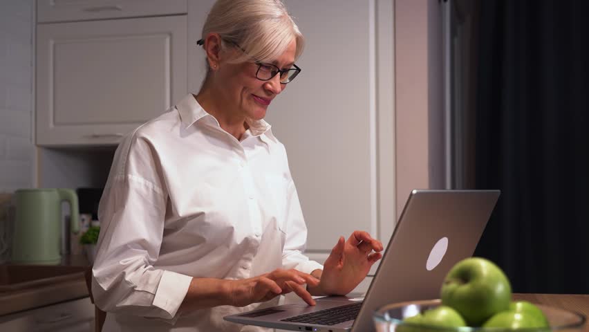 An elderly woman enjoys a virtual call with loved ones on her laptop. She smiles warmly, cherishing the ability to stay connected through online communication and modern technology.