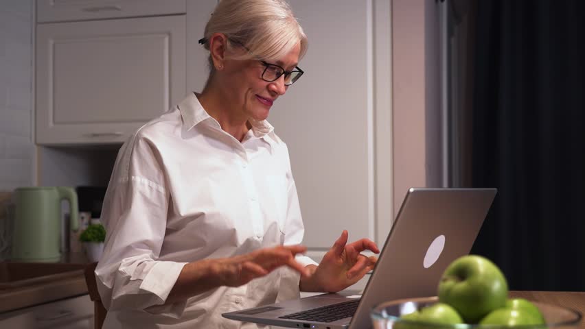 A cheerful senior woman waves at her family during a video call, smiling warmly as she stays connected through digital technology. She enjoys the interaction and feels close to loved ones.