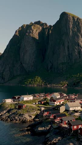 Charming village showcases traditional red cabins along the coastline, directly beneath towering mountains. The serene waters reflect the beauty of nature, Lofoten Norway