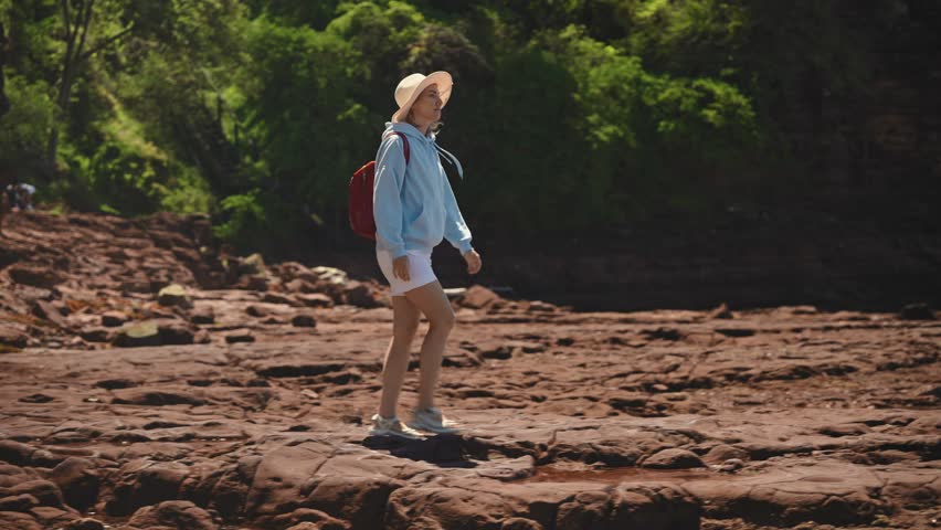 A young, cheerful female tourist wearing a hat and red backpack walks along rocky shoreline toward the water. Stunning Australian coastal landscape with dramatic cliffs and ocean views