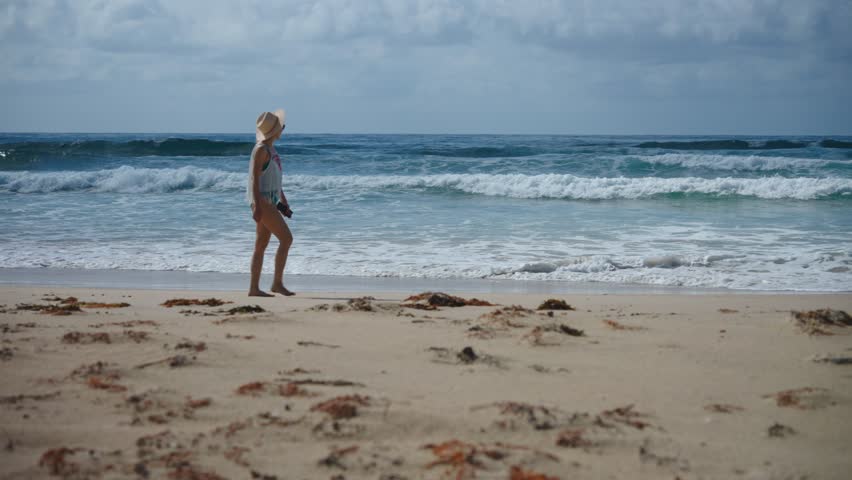 A woman strolls along a sandy beach, her bare feet sinking into the soft shore as waves crash gently in the background. Dressed in a fringed beach cover-up and a straw hat
