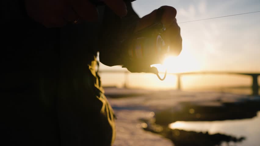 Winter fishing capturing golden sunrise moment, standing on frozen lake with fishing rod, sunlight reflecting on reel and fishing line during peaceful morning activity