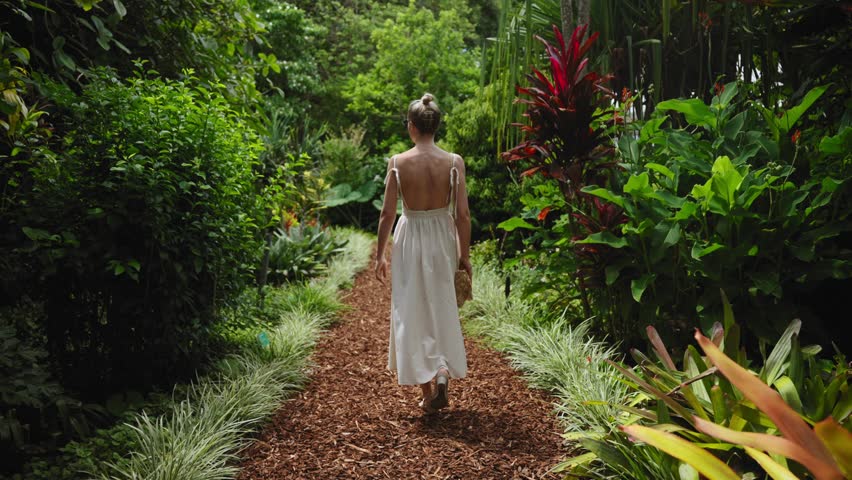 A young, beautiful tourist woman walks through Sydney's Royal Botanic Garden on a bright summer day. Lush greenery, exotic flowers and tropical plants surround her in this vibrant nature setting.