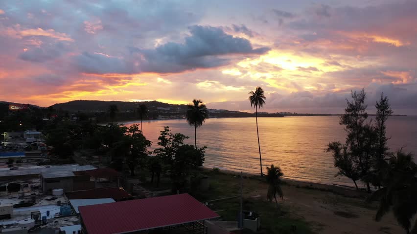 A drone flying on a beach town with tropical greenery, showcasing the colorful sky of sunset reflecting on tranquil sea waters in Luquillo Puerto Rico