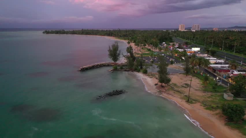 A drone view of Luquillo Beach with clear water, greenery, breakwaters and buildings under colorful sky at sunset in Puerto Rico