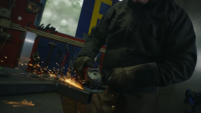 Male welder handling portable professional angle grinder to grind metal in a workshop, medium close up shot. Labor, work, and manufacturing concepts.