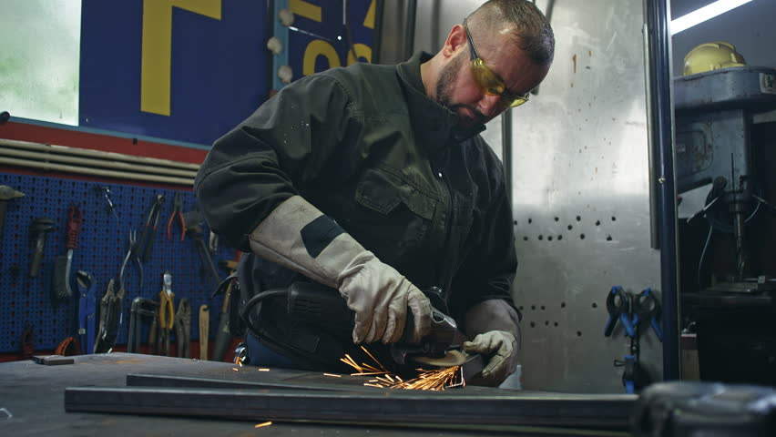 Skilled manual worker wearing protective equipment grinding steel in a welding workshop, handheld shot. Metalwork industry career concept.