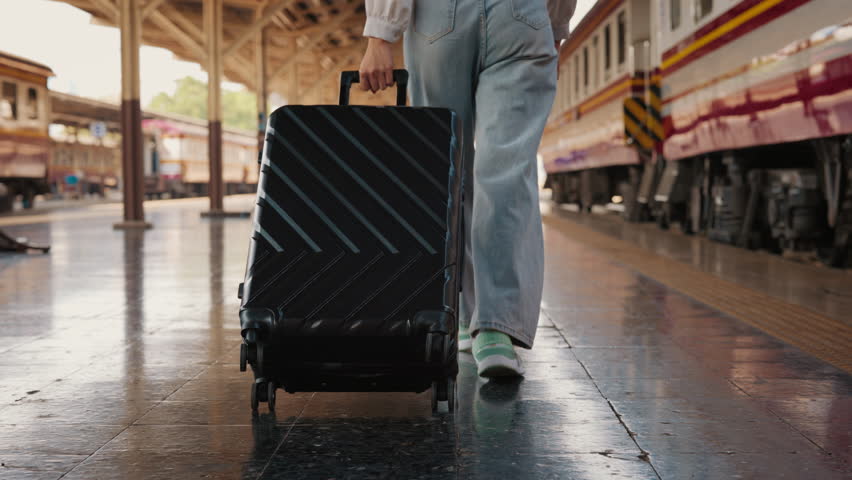 Woman passenger in jeans legs pulling suitcase along railroad platform closeup. Female traveler transports luggage bag for journey at train station backside view. Holiday trip