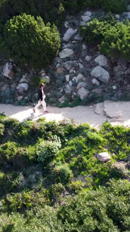 A vertical, ascending drone footage of a woman walking on the shore of Costa Paradiso in Sassari, Sardinia, Italy on a sunny day